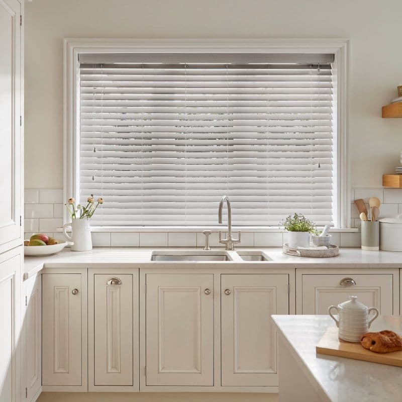 White horizontal venetian blinds covering a large window, slats mostly closed and slightly tilted to filter soft daylight; above a neutral, well-lit kitchen sink with marble countertop, chrome faucet, plants.