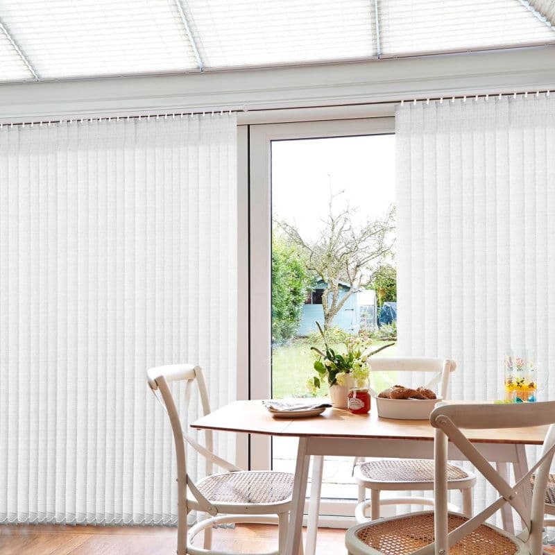 White vertical fabric blinds span floor-to-ceiling sliding doors, sides closed while center panels are parted to reveal light; filtering daylight into a dining area with table, chairs, and garden view.