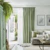Green pinch-pleated curtains with an inner white sheer, partly drawn and filtering daylight beside an open white door. Bright, plant-filled living room with pale sofa, cushions, wooden coffee table, botanical wall art.
L'ART DES FLEURS
