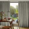 Full-length pleated grey patterned curtains, drawn aside to reveal a patio door, filtering soft daylight; a wooden dining table set with pancakes, fruit and pendant lamp, garden and shed beyond.