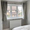Grey pleated, eyelet-top curtains on a metal rod, pulled back with matching tiebacks, framing a three‑pane casement window and filtering daylight into a neutral bedroom with patterned wallpaper and white radiator.