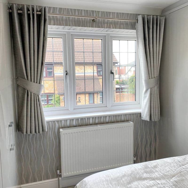 Grey pleated, eyelet-top curtains on a metal rod, pulled back with matching tiebacks, framing a three‑pane casement window and filtering daylight into a neutral bedroom with patterned wallpaper and white radiator.
