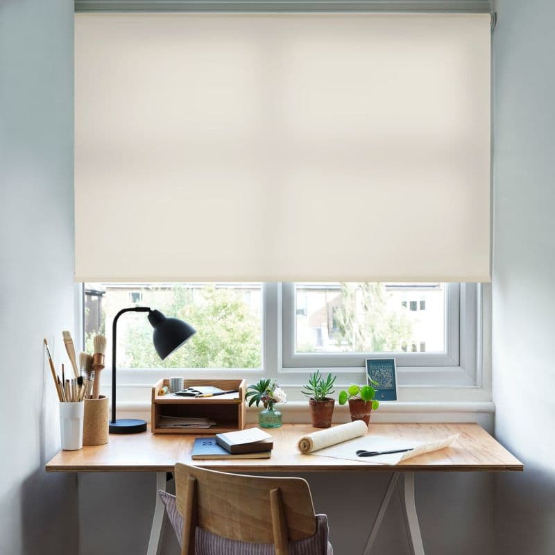 A cream roller blind lowered over a double window, filtering soft daylight; a tidy wooden desk beneath holds a black desk lamp, paintbrushes, small potted succulents, notebooks and a chair.