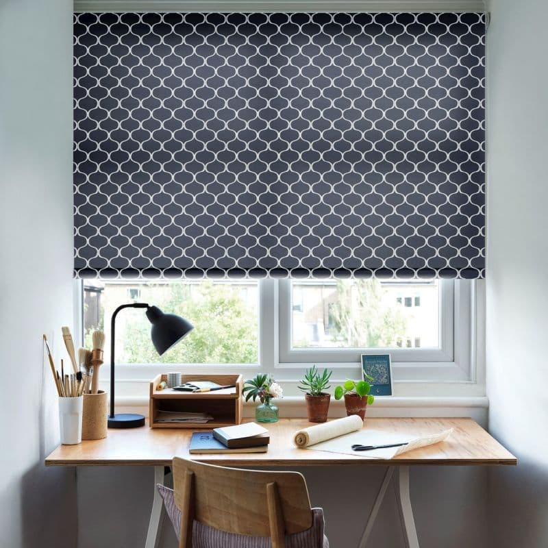 A navy roller blind with a white trellis pattern, pulled down across the window, filtering daylight and obscuring view; below, a light-wood desk with lamp, brushes, notebooks and potted plants.