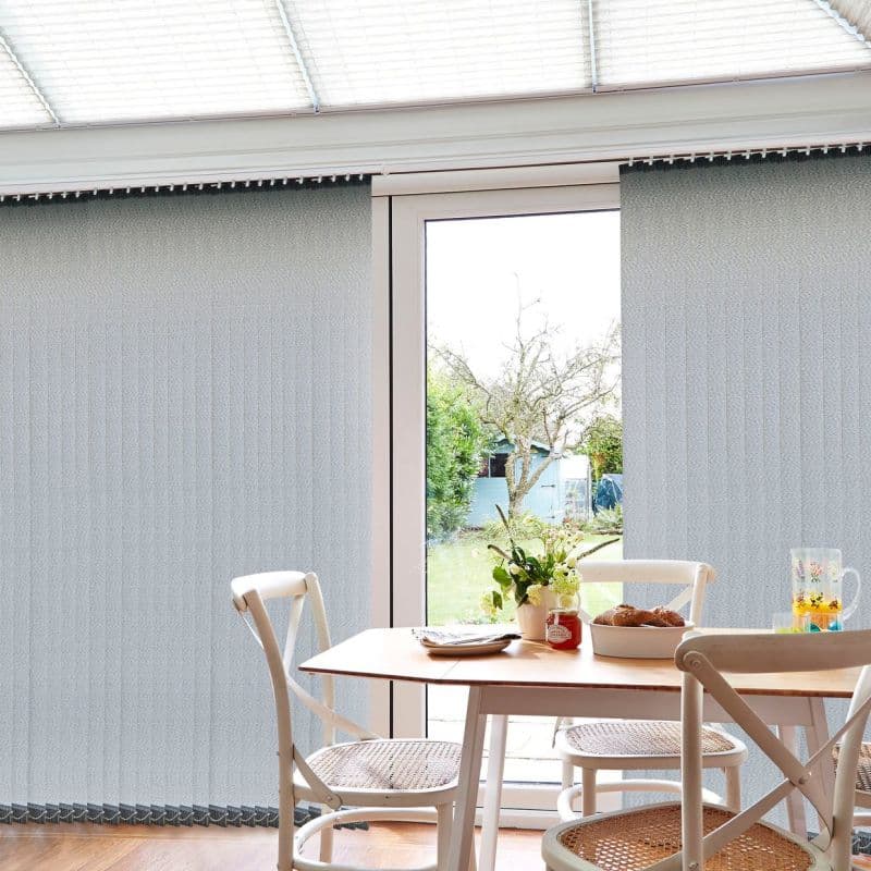Vertical textured grey vertical blinds spanning a sliding glass door, mostly closed with a central opening, filtering daylight; small dining table and chairs in a sunroom, garden visible outside.