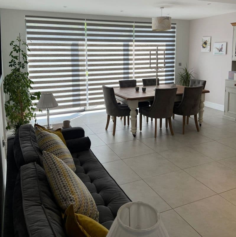 Zebra-style dual roller blinds cover a full-width floor-to-ceiling window, alternating opaque and sheer horizontal stripes filtering daylight into a bright tiled open-plan dining and living area with table and sofa.