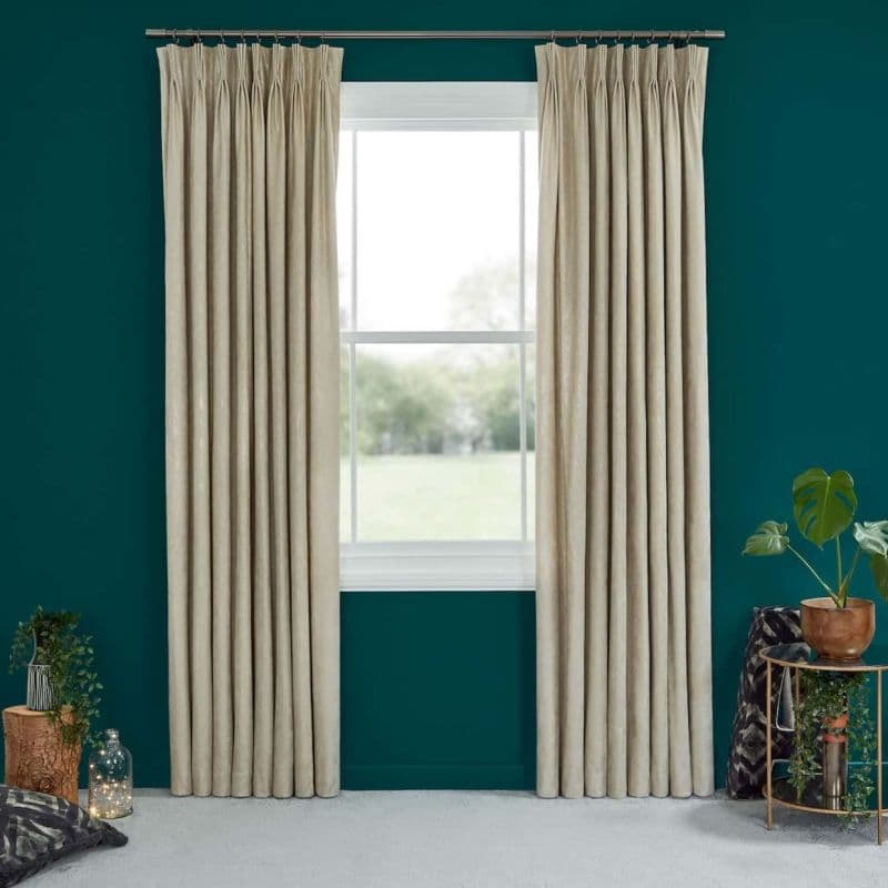 Beige pinch-pleat curtains on a metal rod, drawn to either side, framing and partially revealing a sunlit white-framed window; teal living-room wall, carpeted floor, plants and a side table.