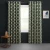 Grommet-top cream curtains with dark green interlocking geometric pattern, hung on a metal rod, parted to frame a daylight window; dark-walled room with a wooden side table and abstract art.