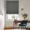 Gray fabric Roman blind lowered over a window, blocking and softly filtering daylight; beneath, a desk with mirror, vase of flowers and teal-upholstered chair in a bright, neutral room.
