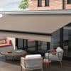 Retractable beige fabric awning extended from the house, supported by a black metal bar, shading a ground-floor patio; modern seating, low wooden table, sliding glass doors, brick upper wall, soft daylight.