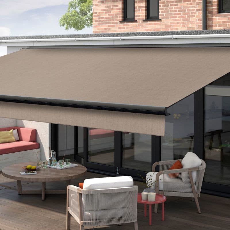 Retractable beige fabric awning extended from the house, supported by a black metal bar, shading a ground-floor patio; modern seating, low wooden table, sliding glass doors, brick upper wall, soft daylight.