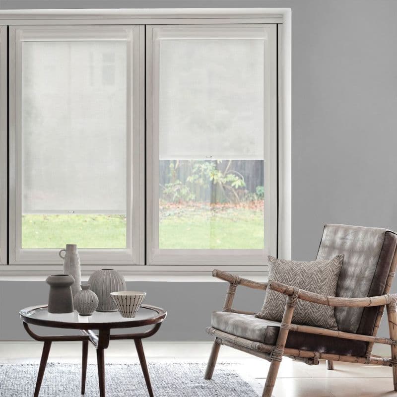 Two white translucent roller blinds—left lowered, right partially raised—softly filter daylight and obscure a backyard; framed by a gray living room with a wooden armchair and small table.