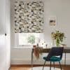 A patterned Roman blind (geometric beige, ochre, gray, black blocks) lowered over a sash window, gently filtering daylight — a bright home office desk below with teal chair and vase of flowers.