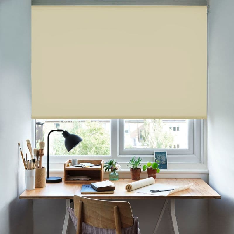 Beige roller blind, lowered over the window with smooth, light‑filtering fabric and hem, filtering daylight and obscuring the view above a wooden desk with lamp, plants, and art supplies.