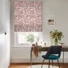 Roman-style fabric blind, white with red maze-like pattern, lowered over the sash window; filtering daylight. Context: small home desk and teal chair, mirror, vase of flowers, and framed seaside print.