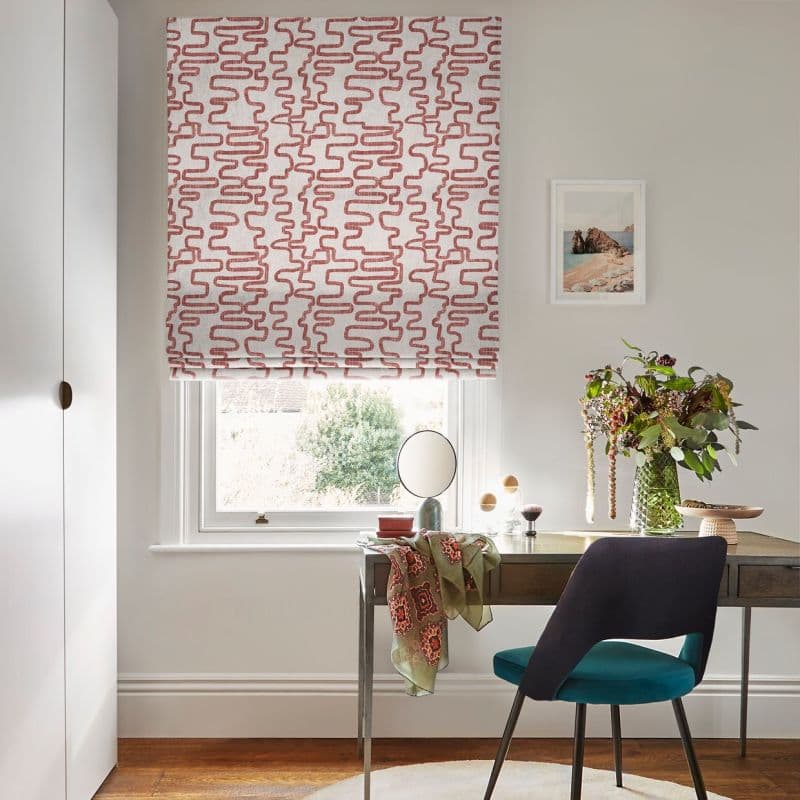 Roman-style fabric blind, white with red maze-like pattern, lowered over the sash window; filtering daylight. Context: small home desk and teal chair, mirror, vase of flowers, and framed seaside print.