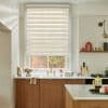 A beige dual-layer zebra roller blind, lowered with alternating sheer and opaque stripes, partially filtering daylight over a kitchen sink with brass faucet, marble countertop, wood cabinets and brick view.