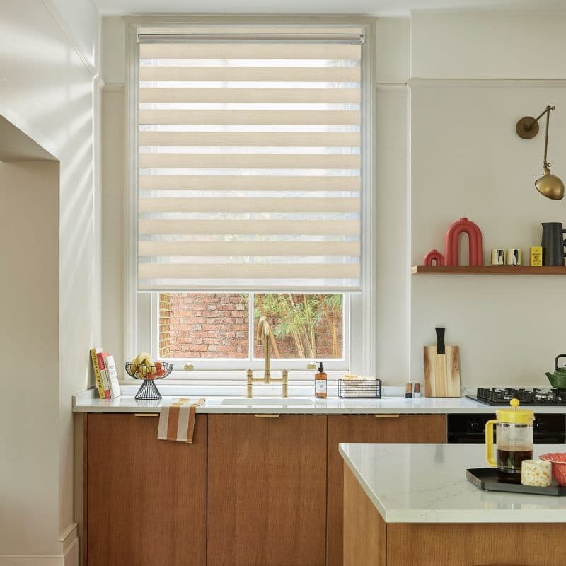 A beige dual-layer zebra roller blind, lowered with alternating sheer and opaque stripes, partially filtering daylight over a kitchen sink with brass faucet, marble countertop, wood cabinets and brick view.