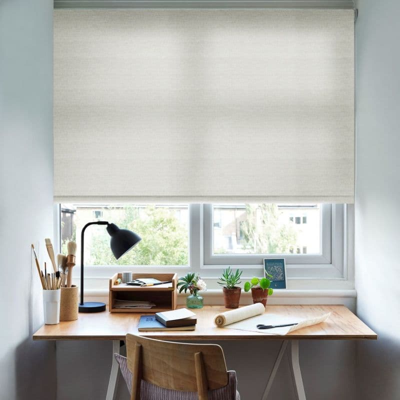 Cream textured roller blind, lowered across the window with a narrow bottom gap; diffusing daylight over a tidy wooden desk workspace with lamp, brushes, plants, and notepads.
