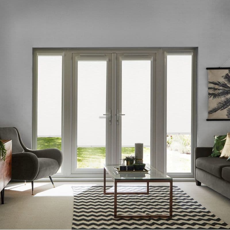 White roller blinds lowered over four tall panes (central French doors with sidelights), filtering daylight, blocking view; living-room grey armchair, sofa, coffee table on patterned rug, lawn glimpsed below.