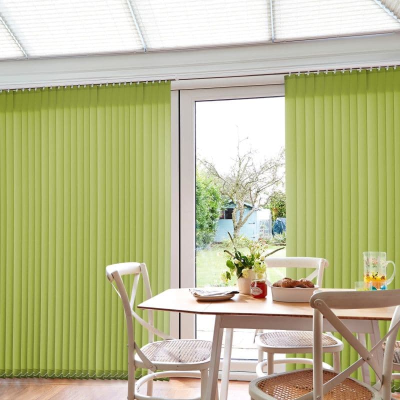 Lime-green vertical blinds, mostly closed with a central gap, filtering daylight through patio doors; small dining table with woven-seat chairs in a sunlit breakfast nook, garden and shed visible outside.