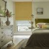 Yellow Roman blind lowered over a window with a patterned sheer beneath, filtering daylight into a cozy bedroom with an olive‑green upholstered headboard, bedside table, floral‑painted dresser, rug, and pendant light.