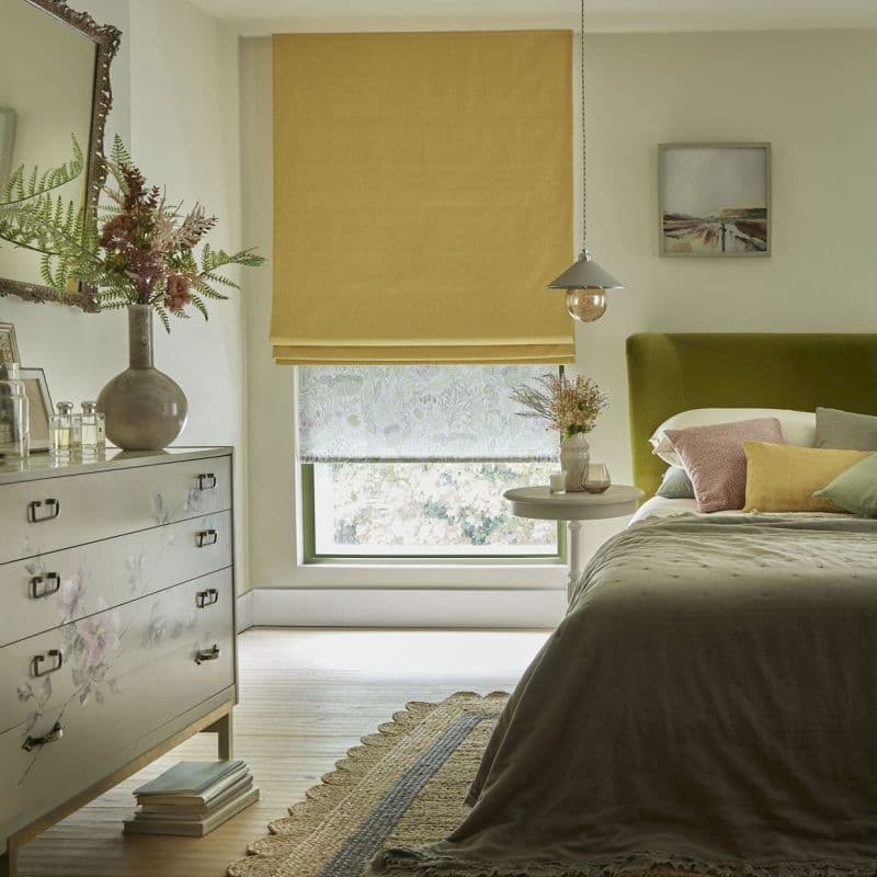 Yellow Roman blind lowered over a window with a patterned sheer beneath, filtering daylight into a cozy bedroom with an olive‑green upholstered headboard, bedside table, floral‑painted dresser, rug, and pendant light.