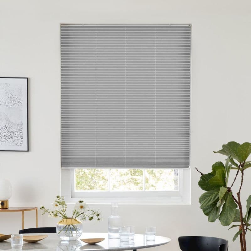 Light-gray pleated cellular blind, fully lowered across the window with subtle horizontal ribs, filtering daylight; set in a white-walled dining area with table, vase of flowers and a potted plant.