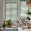 Horizontal pale-green Venetian blinds on a door and adjacent window, closed and softly filtering daylight; kitchen scene with small tabletop Christmas trees, gingerbread houses, candy canes and baking tools.