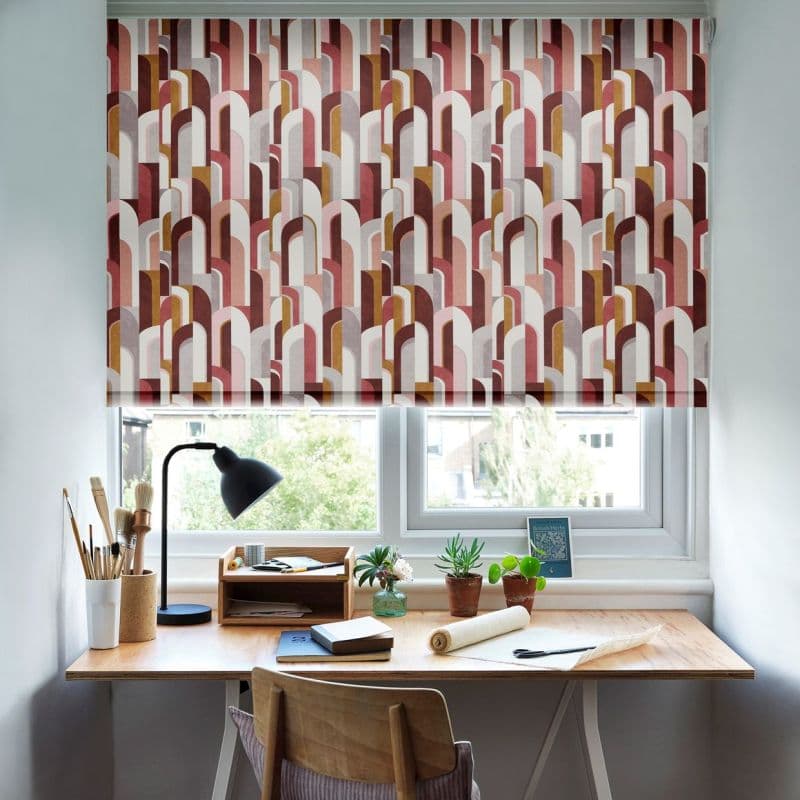 Patterned roller blind with multicolored geometric arches, lowered and filtering daylight; set over a window above a wooden desk with lamp, plants and stationery in a small home office.