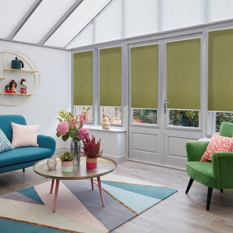 Olive-green roller blinds lowered across French doors and tall windows, partially blocking sunlight; set in a bright conservatory-style living room with skylight, teal sofa, pastel rug and plants.