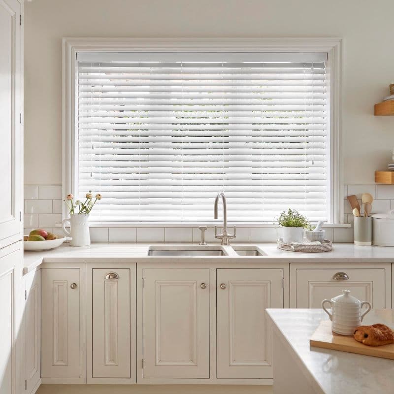 White horizontal blinds covering a large window, lowered with slats mostly closed, filtering soft daylight; above a farmhouse-style sink in a bright, neutral kitchen with countertops and utensils.