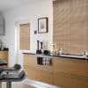 Beige horizontal Venetian blinds covering two windows, fully lowered and closed, filtering soft daylight; foreground oak kitchen cabinets and black countertop with utensils, glass jars, and two black bar stools.