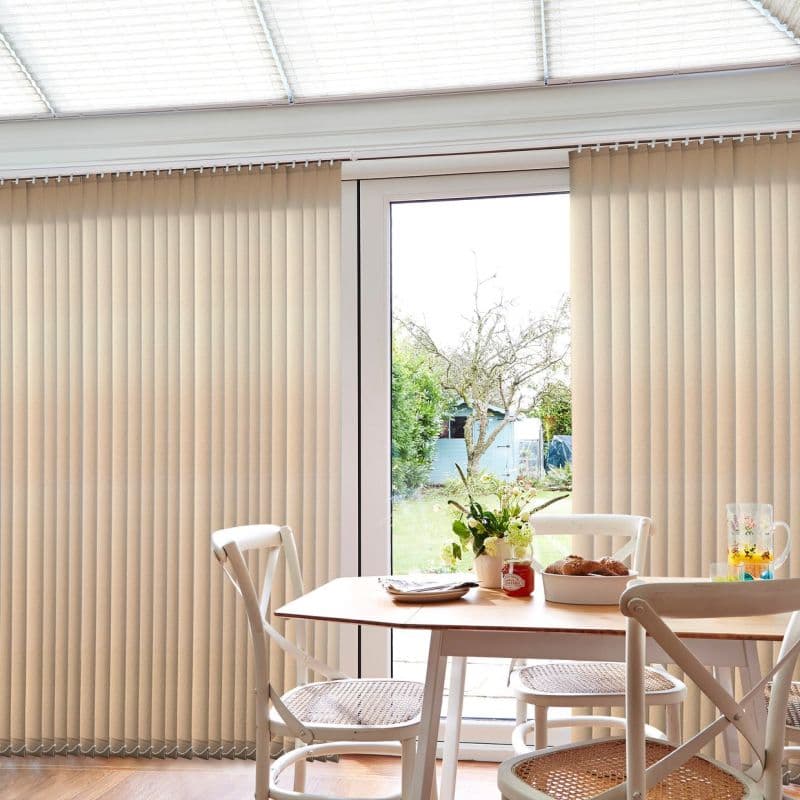 Beige vertical blinds cover a large patio door, mostly closed with a central gap revealing glass; they filter daylight into a dining area with table, chairs, vase, and garden view.
