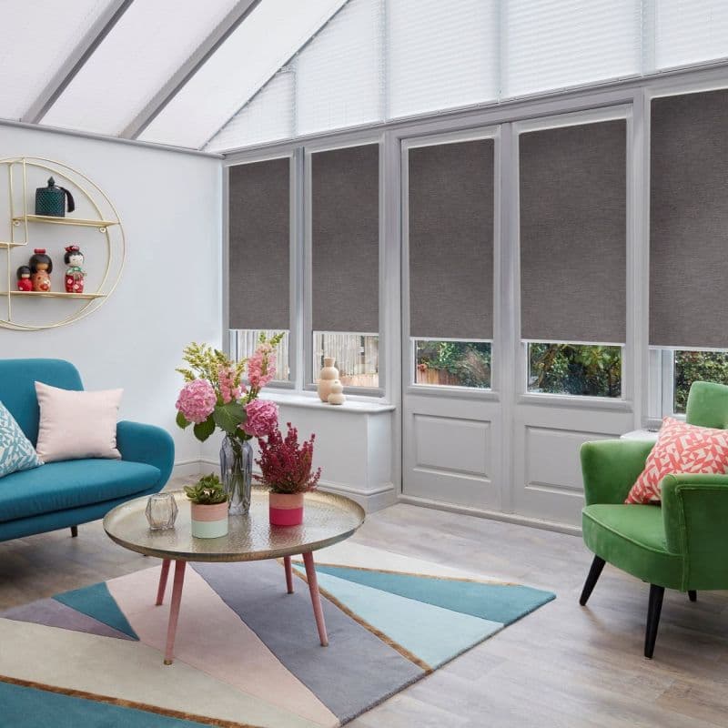 Grey fabric roller blinds covering four tall window panels, lowered to mid-height leaving lower glass clear; filtering daylight into a conservatory living room with colorful sofas, rug and coffee table.