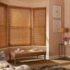 Wooden horizontal Venetian blinds covering a three-panel bay window, closed and filtering warm sunlight into a softly lit living room with upholstered bench, side table, and lamp.