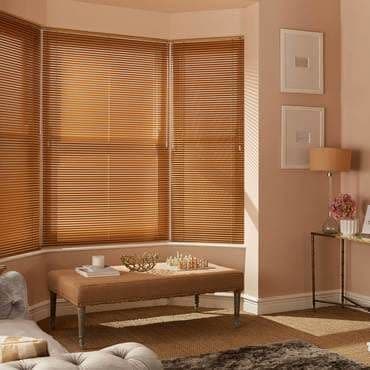 Wooden horizontal Venetian blinds covering a three-panel bay window, closed and filtering warm sunlight into a softly lit living room with upholstered bench, side table, and lamp.