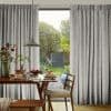 Pleated floor-to-ceiling gray patterned curtains, drawn aside to reveal a glass door; hanging in soft folds. Wooden dining table set for breakfast under a pendant lamp, sunlit garden beyond.