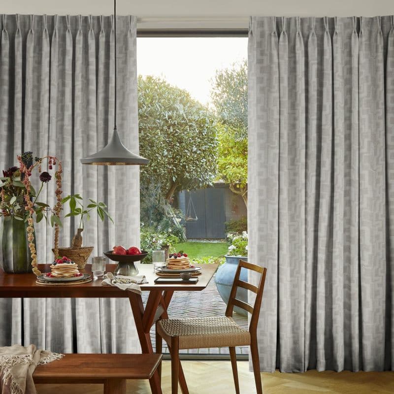 Pleated floor-to-ceiling gray patterned curtains, drawn aside to reveal a glass door; hanging in soft folds. Wooden dining table set for breakfast under a pendant lamp, sunlit garden beyond.