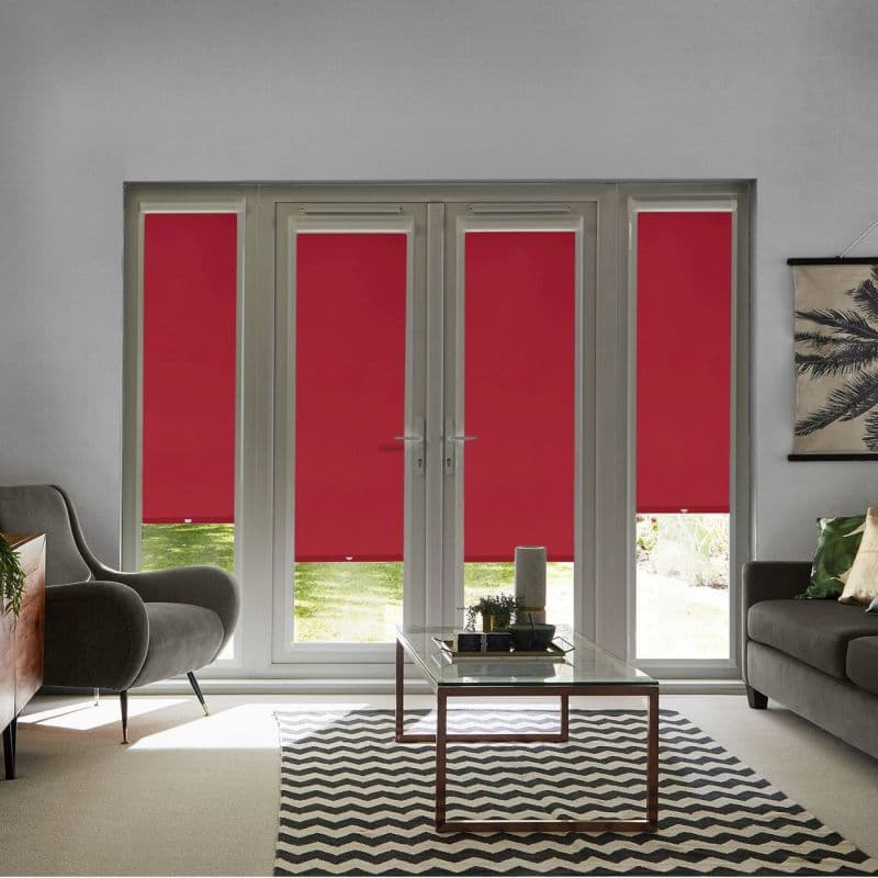 Red roller blinds cover four-panel glass patio doors and sidelights, pulled down to block light; living room with gray sofas, glass coffee table on chevron rug, garden view glimpses.