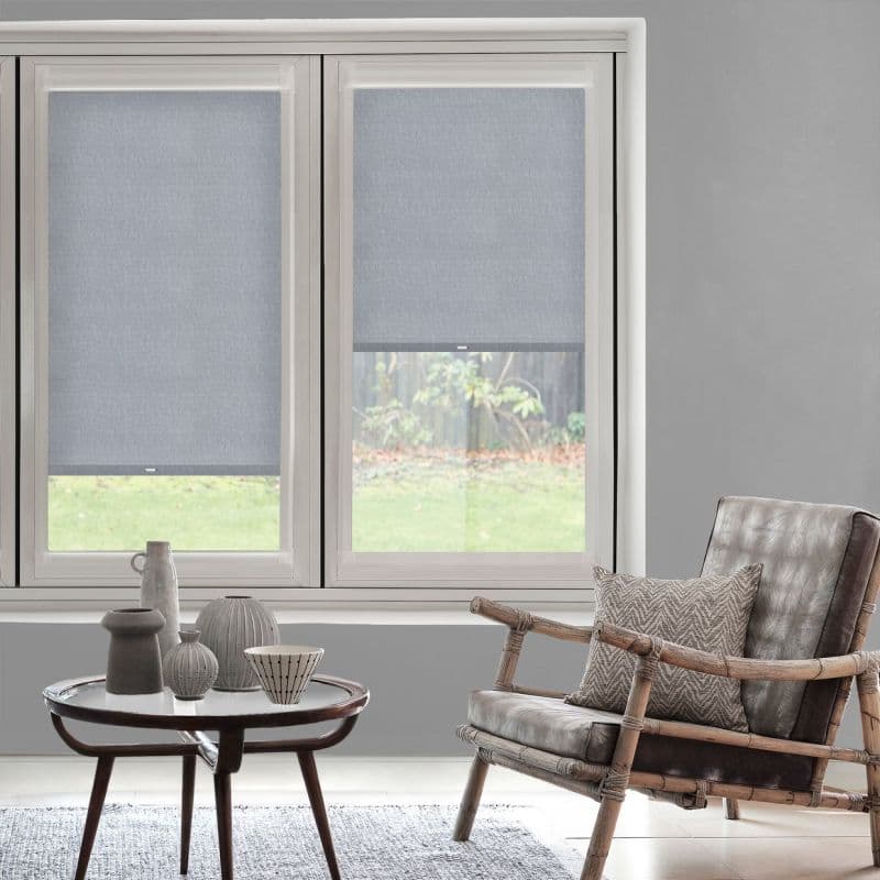 Two light-gray fabric roller blinds covering a double window, partly lowered and filtering daylight; modern living room with neutral walls, rattan armchair with cushion, round coffee table and garden view.