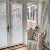 White horizontal Venetian blinds on double French doors and side window, closed with slats tilted to filter light; sunroom corner with wicker chair, floral cushion, radiator, tiled floor, brick exterior.