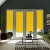 Tall glass French doors fitted with bright yellow roller blinds, blocking light, centered in a gray-walled living room with green armchairs, glass coffee table, chevron rug, and garden beyond.