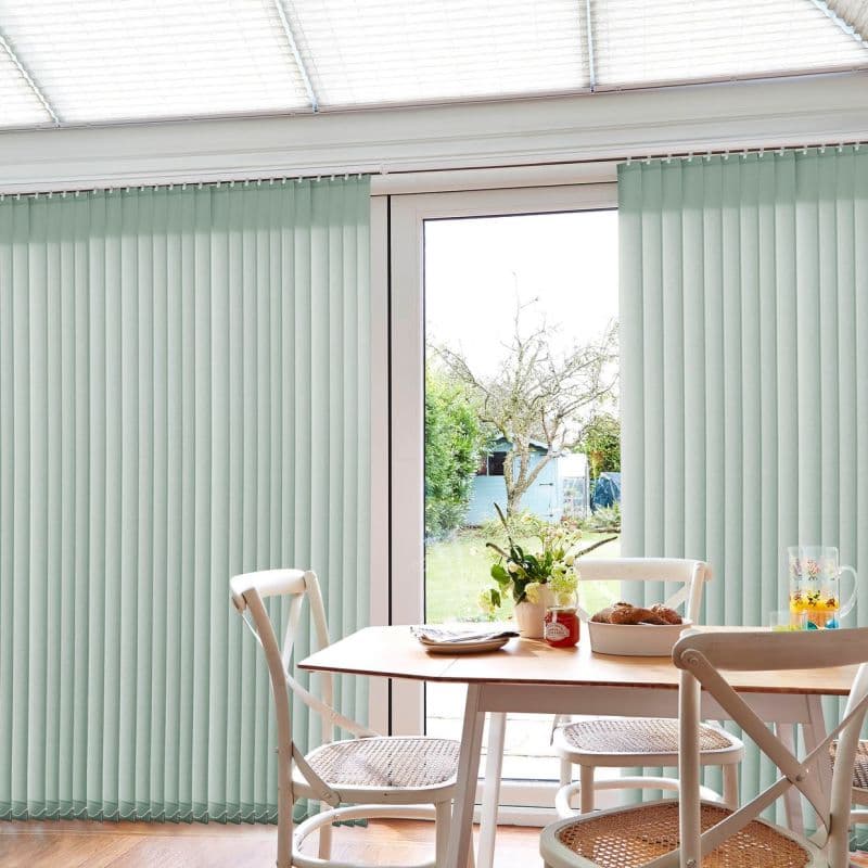 Pale mint vertical blinds span a patio door, slatted and mostly closed with a central opening, filtering soft daylight over a small dining table with chairs and a garden view.