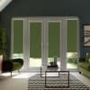 Four glass patio doors with olive-green roller blinds are closed, blocking most view; set in a dark-walled living room with green armchairs, glass coffee table, chevron rug, and plants.