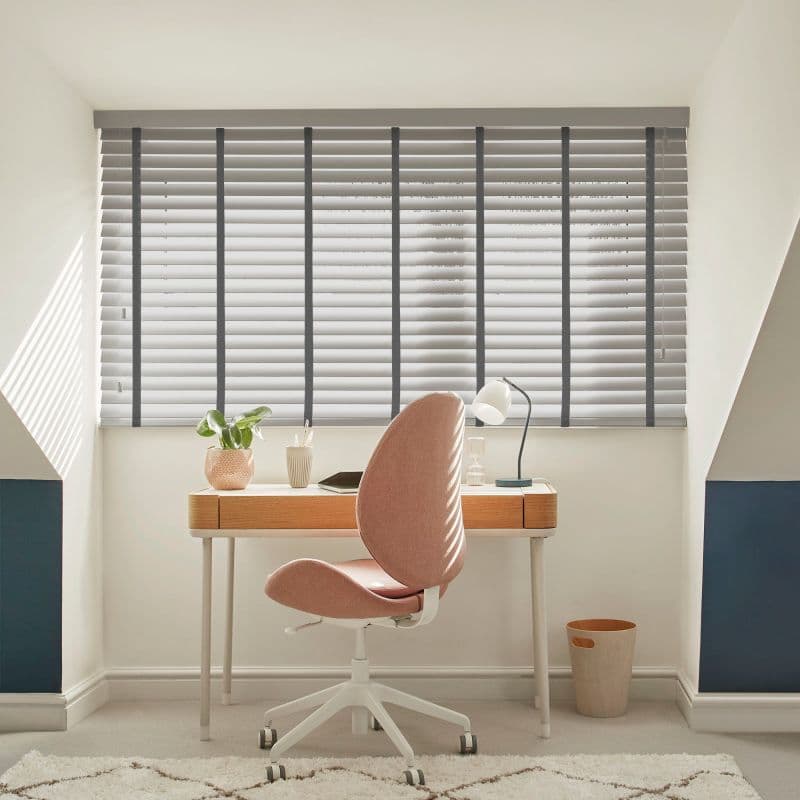 White horizontal blinds with dark vertical tapes covering an alcove window, slats mostly closed and filtering soft daylight, casting striped shadows over a compact home-office desk with pink swivel chair.
