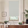 A patterned roller blind covers a window above the sink, partially blocking daylight; countertop holds faucet, bowls of leafy greens, potted plants and framed sketch on kitchen wall.