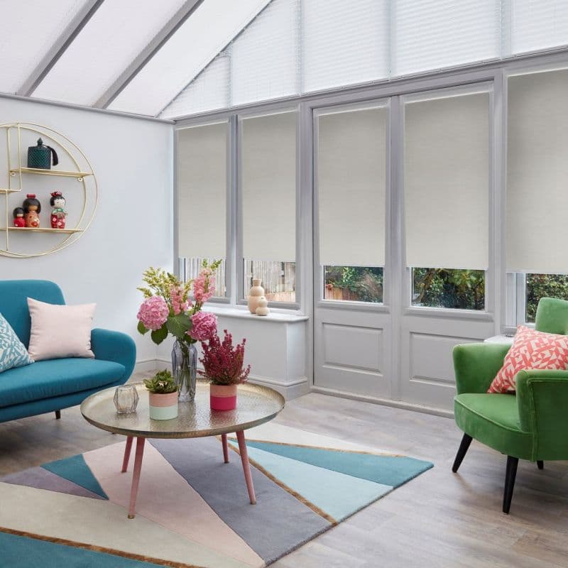 Beige roller blinds, half-lowered over tall glazed doors and windows, filtering daylight; conservatory-style lounge with teal sofa, green armchair, round coffee table and flowers on a pastel geometric rug.