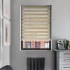 Zebra-style roller blind with alternating beige wood‑grain and sheer horizontal stripes, partially lowered, filtering daylight; set in a grey-walled kitchen nook with a coffee machine, radiator below, and brick building view.