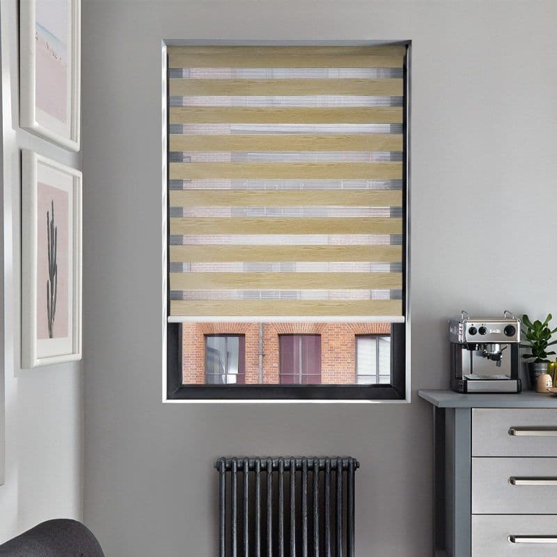 Zebra-style roller blind with alternating beige wood‑grain and sheer horizontal stripes, partially lowered, filtering daylight; set in a grey-walled kitchen nook with a coffee machine, radiator below, and brick building view.
